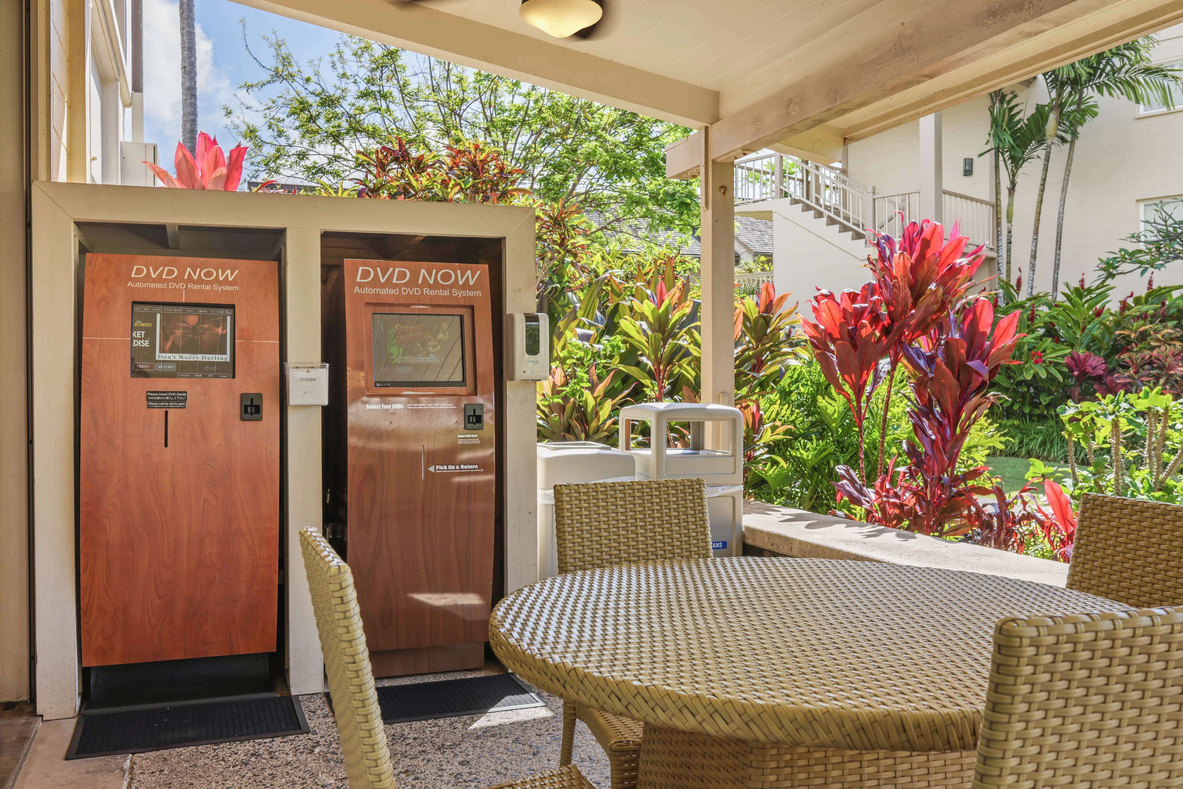 A DVD rental kiosk with a sign, a covered outdoor seating area with wicker furniture, and lush tropical plants.