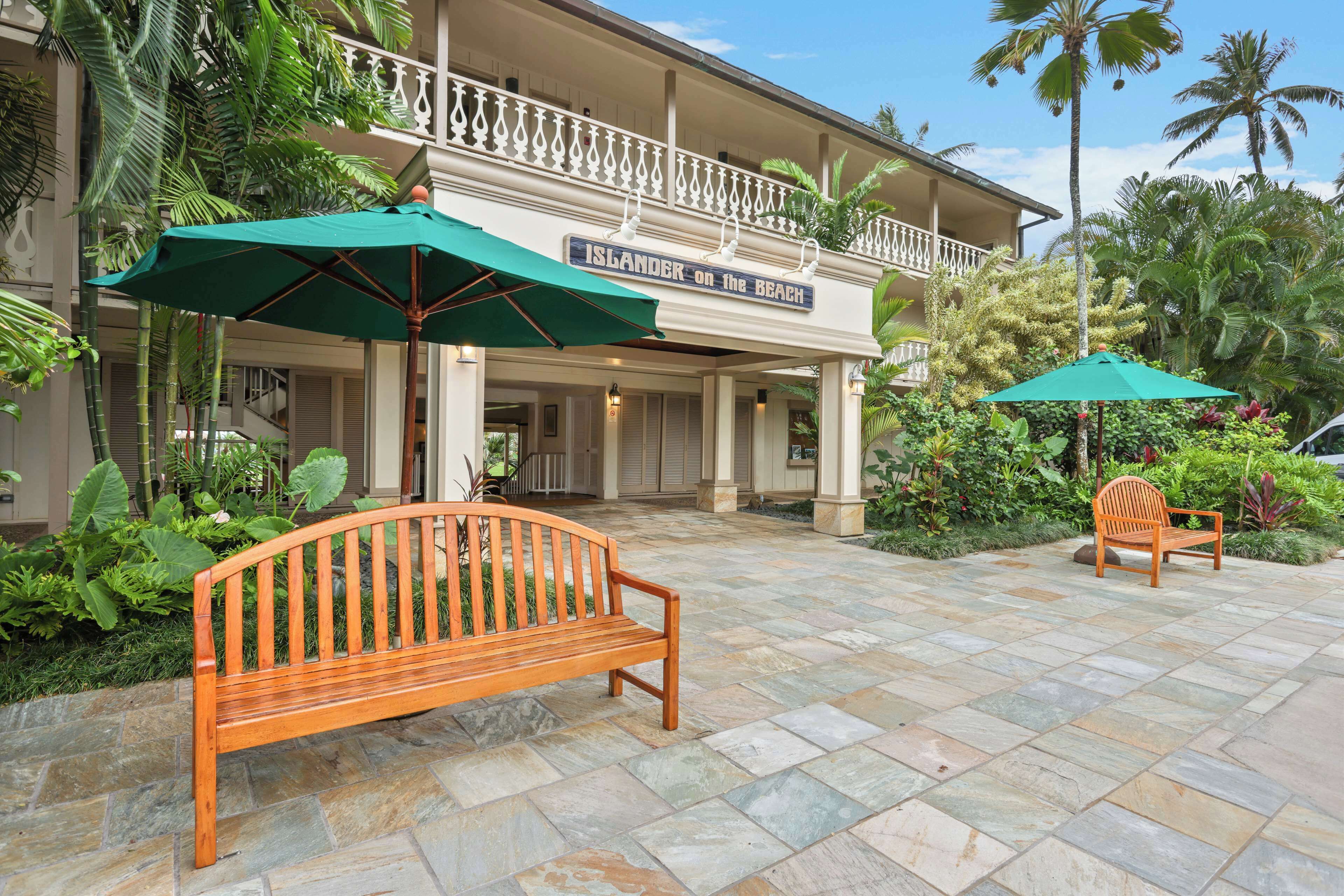 A wooden bench and a chair under a green umbrella in front of a building with a balcony.
