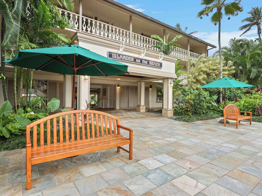 A wooden bench and a chair under a green umbrella in front of a building with a balcony.