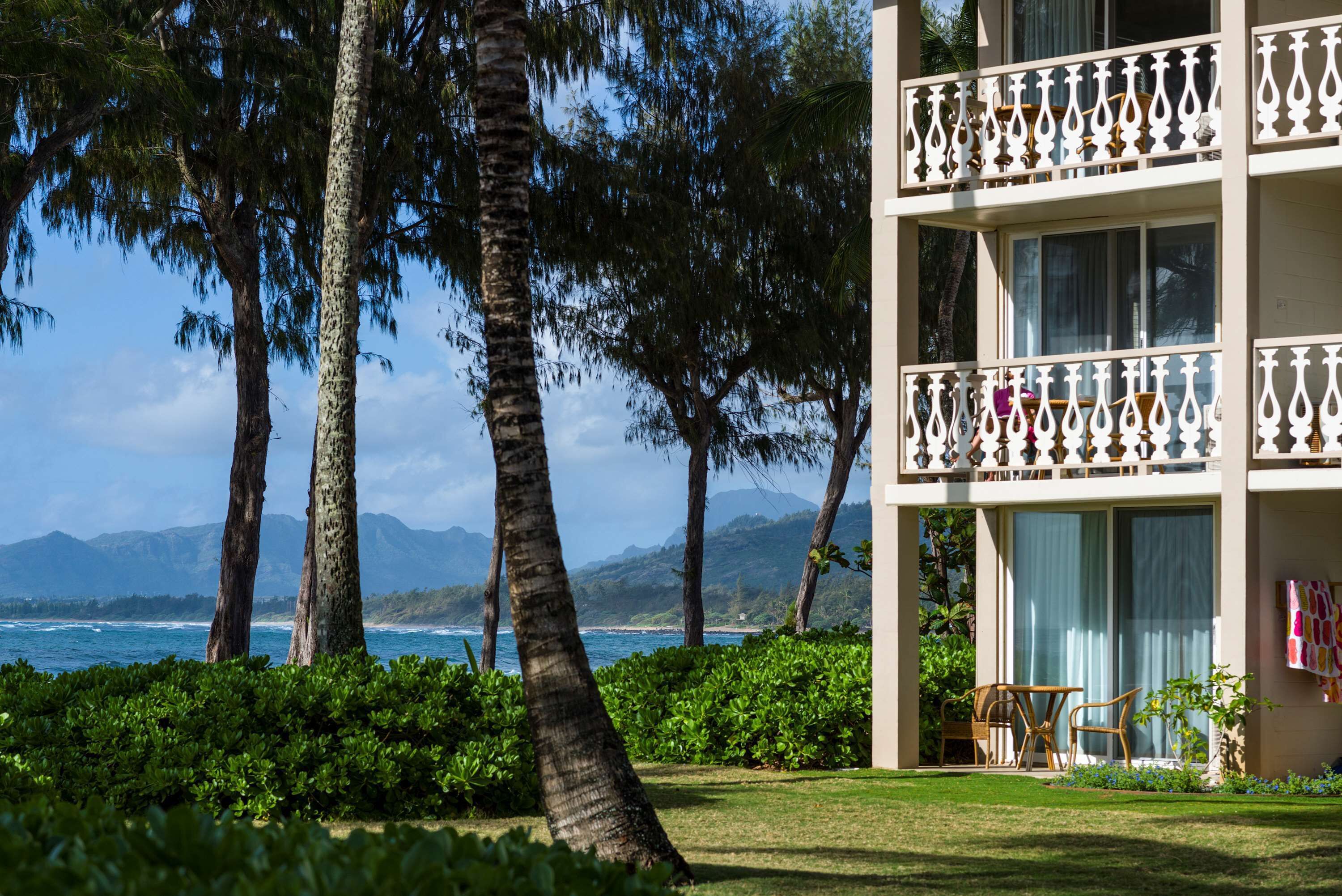 A beachfront property with a balcony, lush greenery, and a view of the ocean and mountains.
