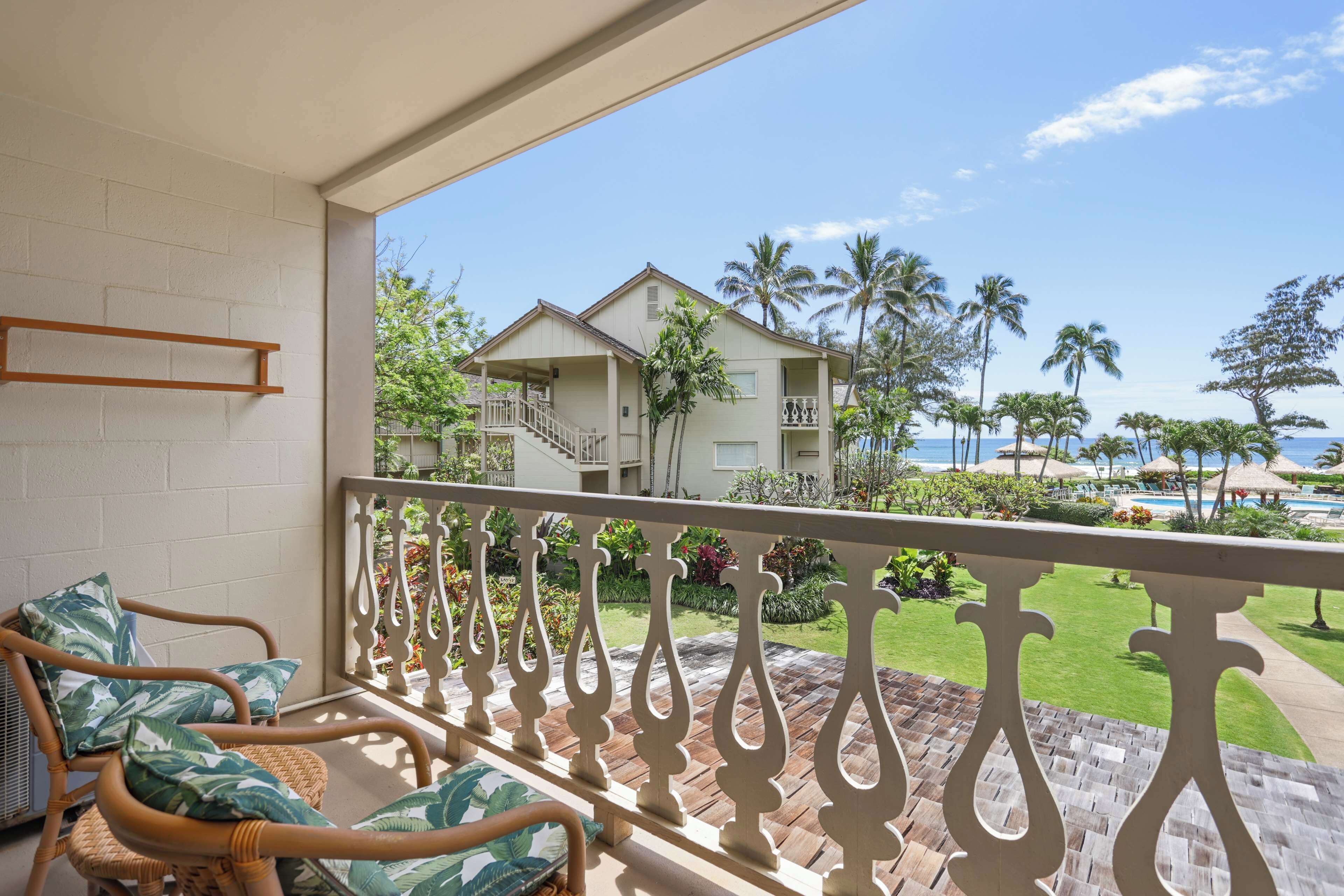 A balcony with wicker chairs and a view of a resort with palm trees and a pool.