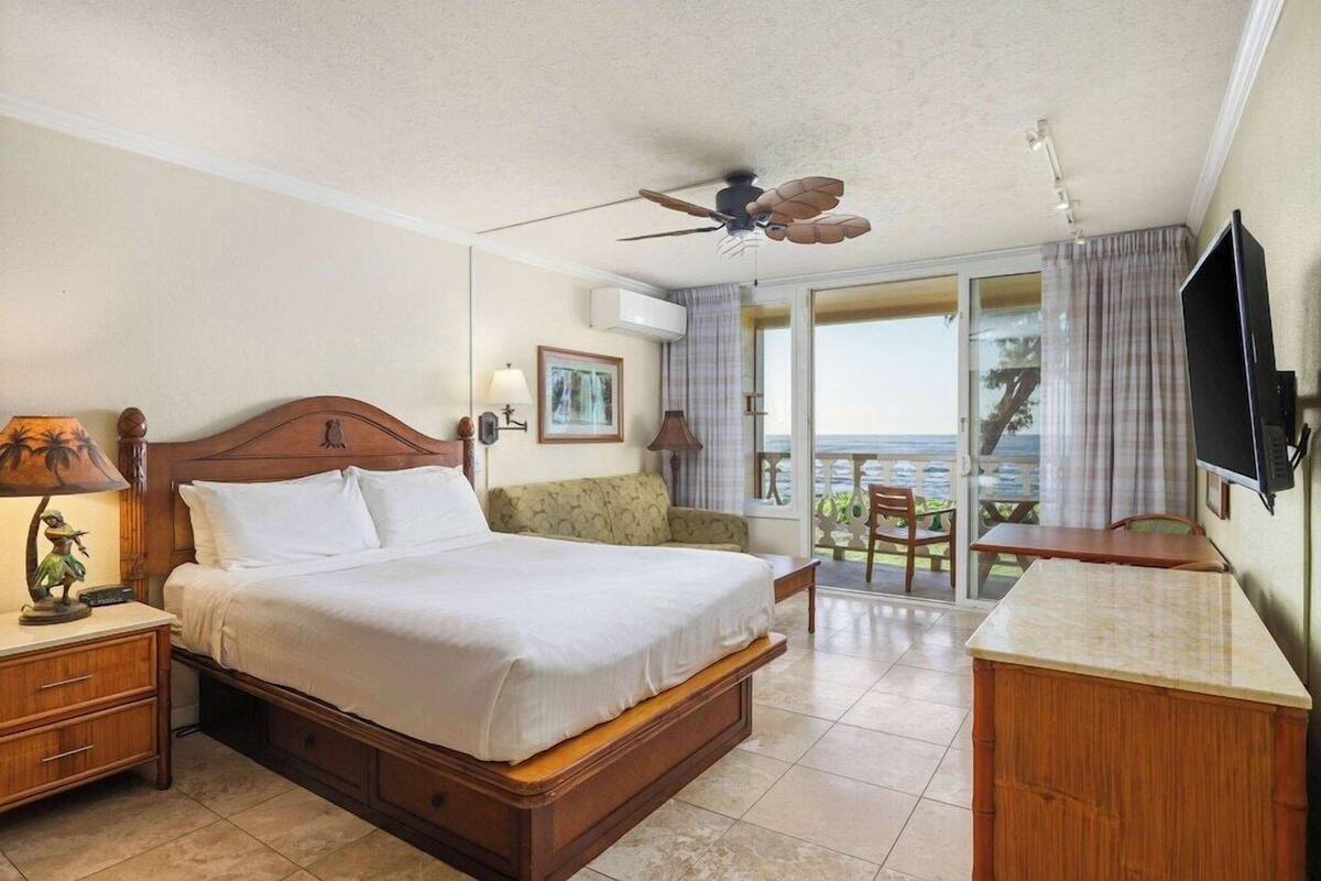 Habitación de hotel con cama, escritorio, televisor, ventilador de techo y balcón con vistas.