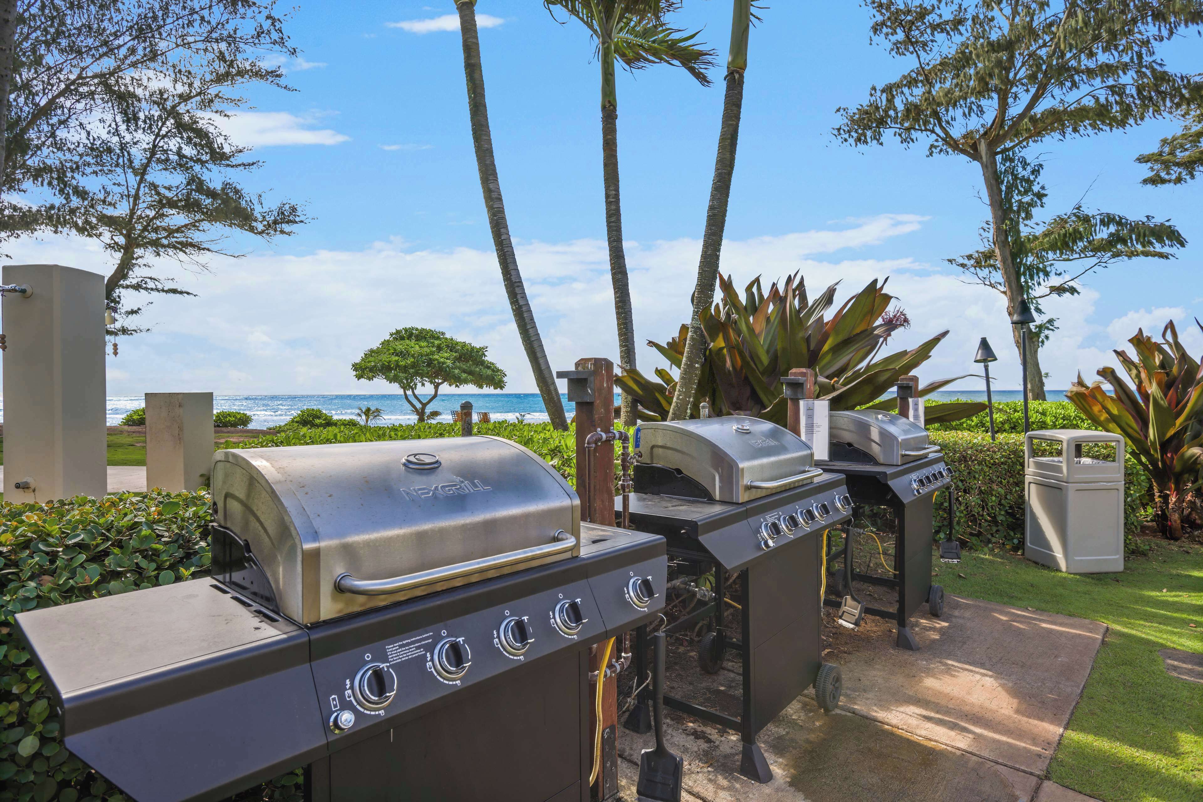 A patio with two gas grills and a trash can, surrounded by greenery and a view of the ocean.