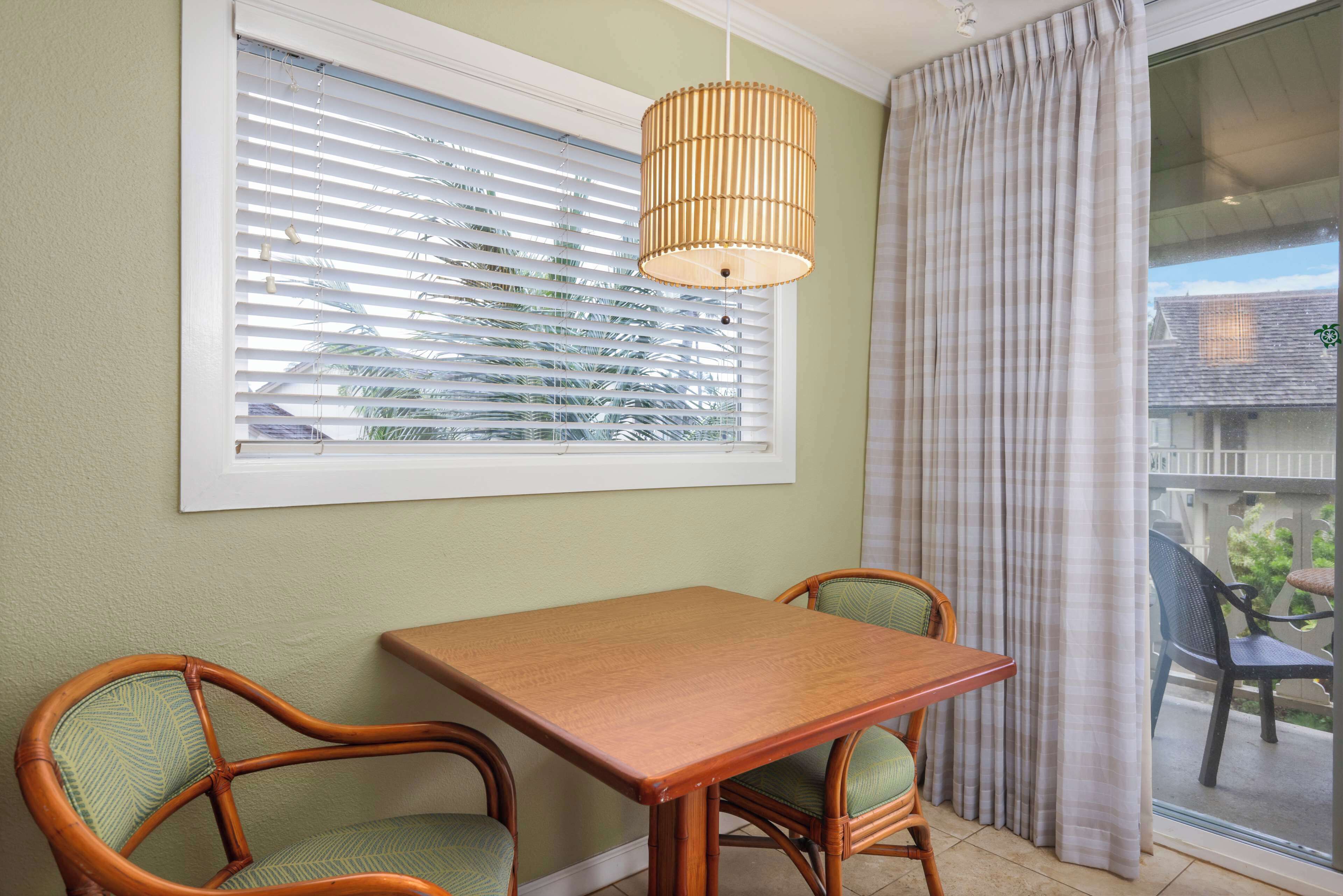 Ein Zimmer mit einem Holztisch und zwei Korbstühlen, einem Fenster mit Jalousien und Blick ins Freie.