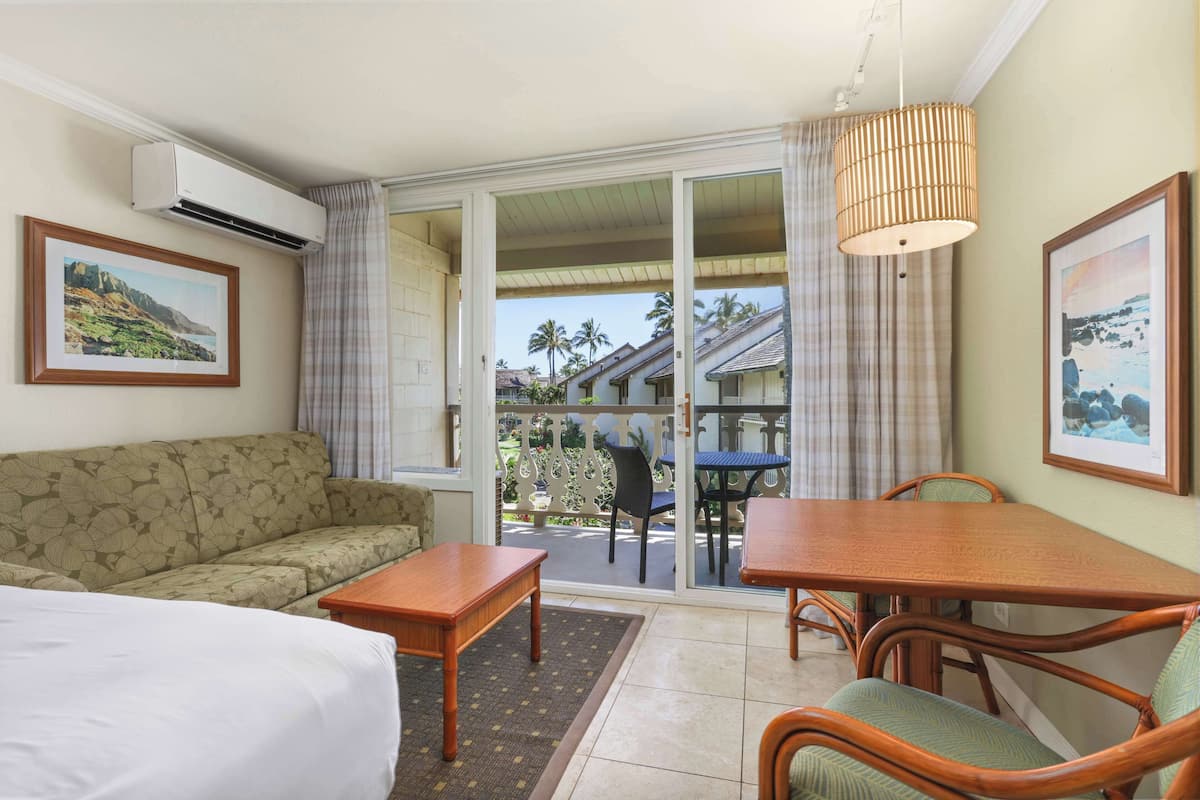 Habitación de hotel con sofá, una mesa de madera y un balcón con vistas a palmeras y un edificio.