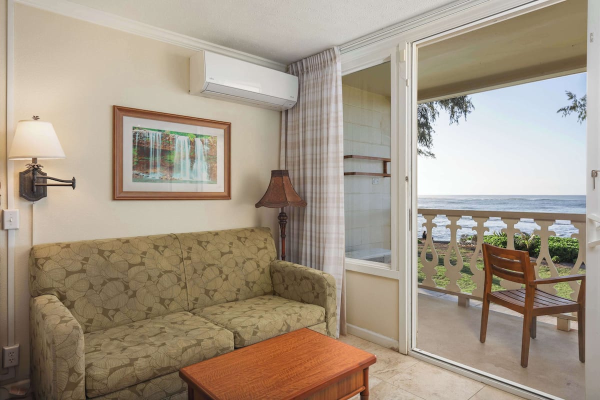 Una sala de estar con un sofá, una mesa de madera, una lámpara, un cuadro enmarcado y vista al mar desde un balcón.