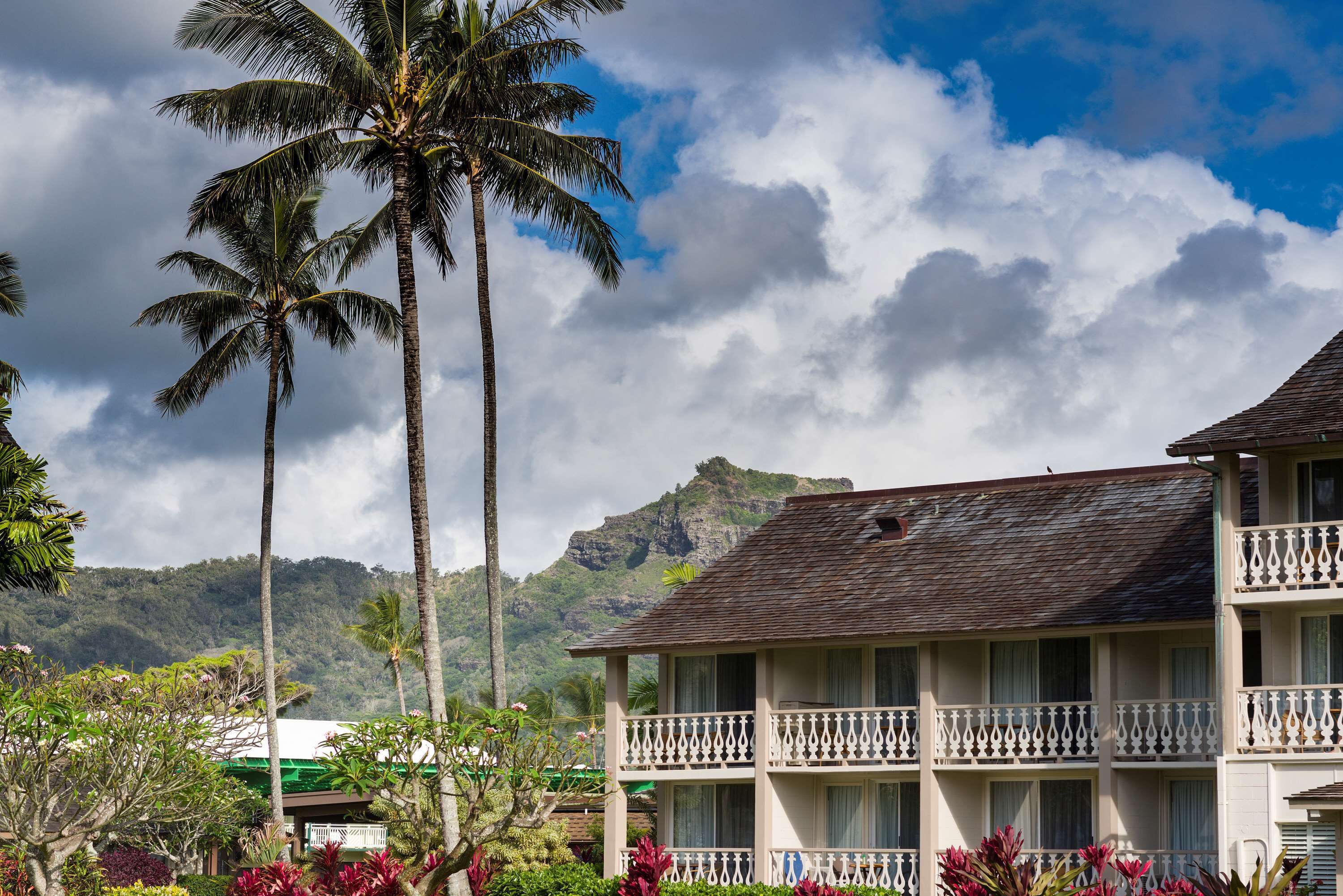 Un hotel con balcón, rodeado de palmeras y abundante vegetación, con una montaña al fondo.