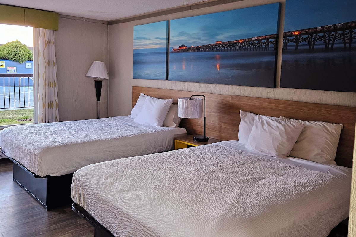A hotel room with two beds, a nightstand, a lamp, and a large painting of a pier at dusk.