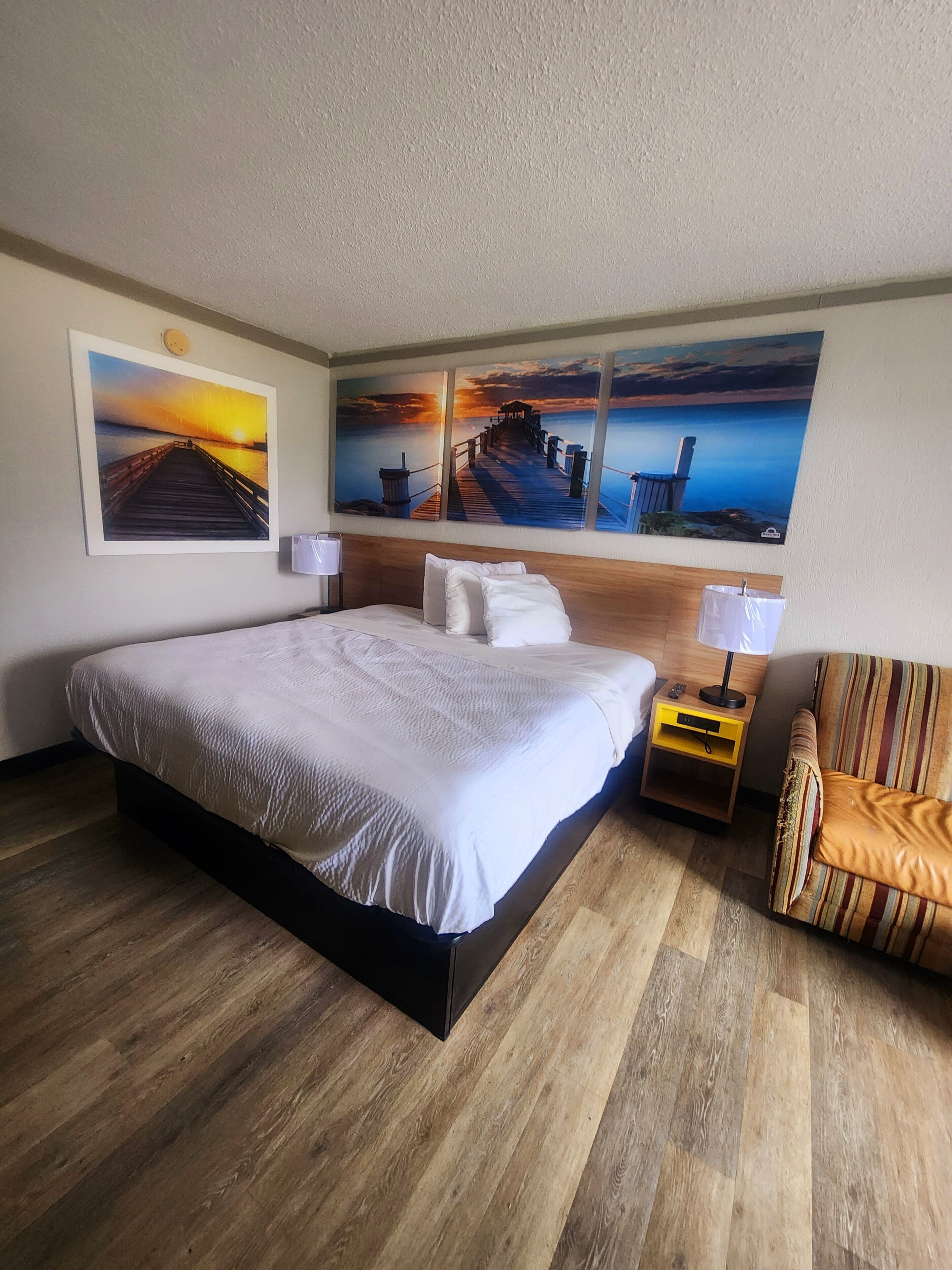 A hotel room with a large bed, wooden flooring, a nightstand with a lamp, a striped chair, and three sunset paintings on the wall.