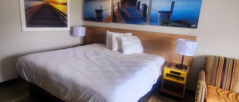 A hotel room with a large bed, wooden flooring, a nightstand with a lamp, a striped chair, and three sunset paintings on the wall.