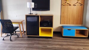 Habitación de hotel pequeña y funcional con escritorio, refrigerador, microondas, televisor y armario con perchas.