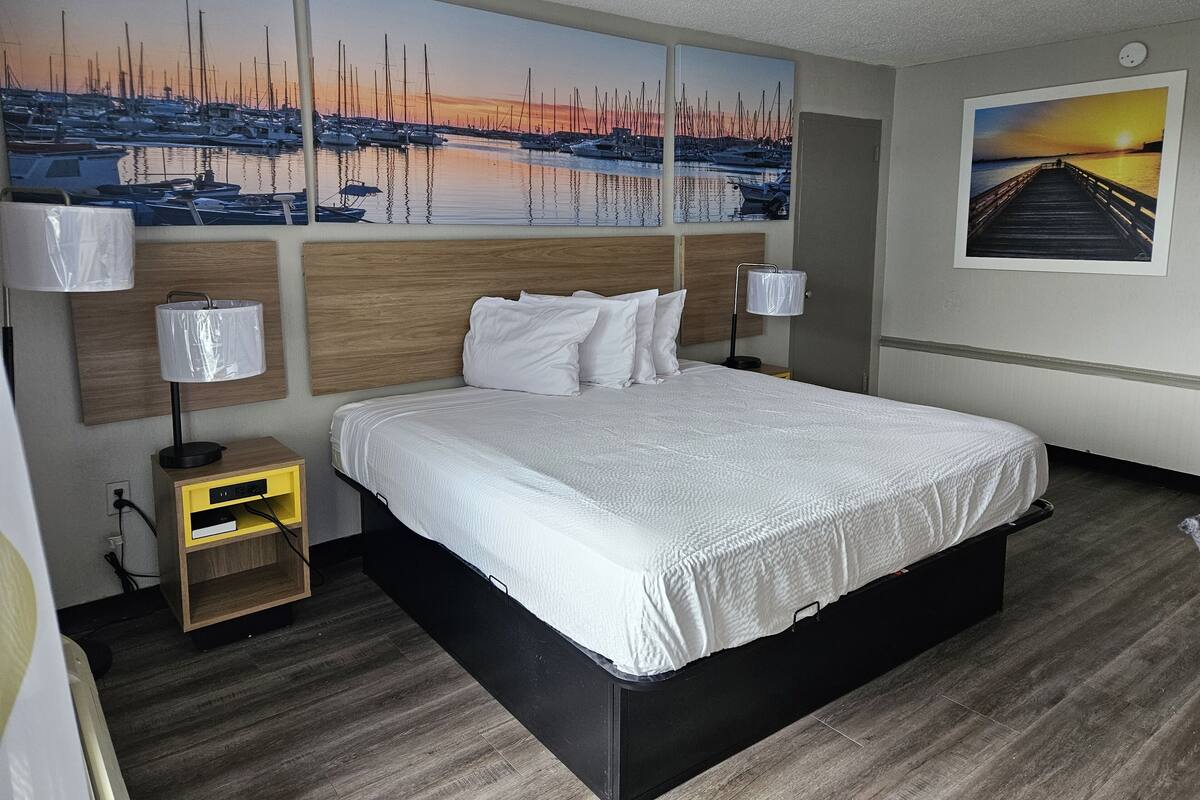 A hotel room with a large bed, wooden headboard, two bedside lamps, a nightstand, and artwork depicting a marina and sunset.