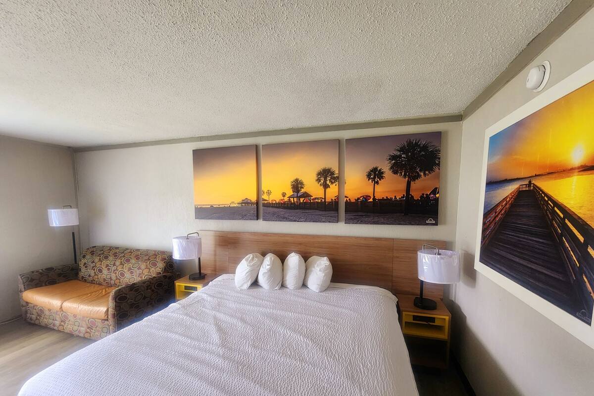 A hotel room with a large bed, two bedside tables, a chair, and three sunset paintings on the wall.