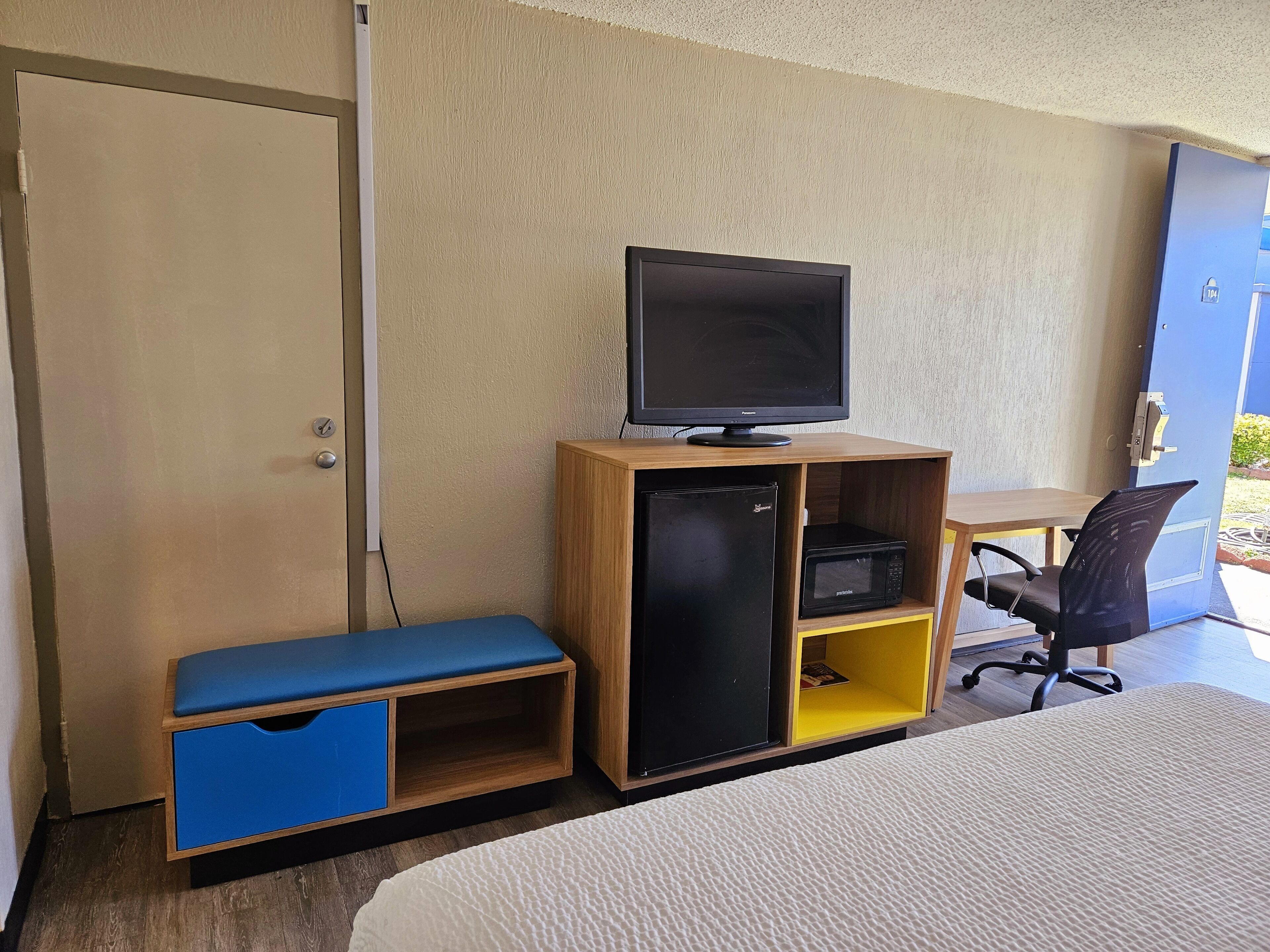 Habitación de hotel con cama, escritorio con monitor de computadora, refrigerador pequeño, microondas, banco de guardado azul y puerta.