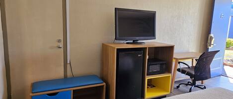 Habitación de hotel con cama, escritorio con monitor de computadora, refrigerador pequeño, microondas, banco de guardado azul y puerta.