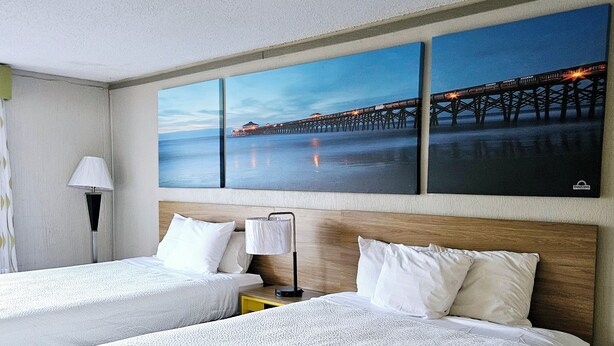 Habitación de hotel con dos camas, una mesita de noche, una lámpara y un cuadro grande de un muelle.