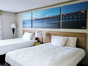Habitación de hotel con dos camas, una mesita de noche, una lámpara y un cuadro grande de un muelle.