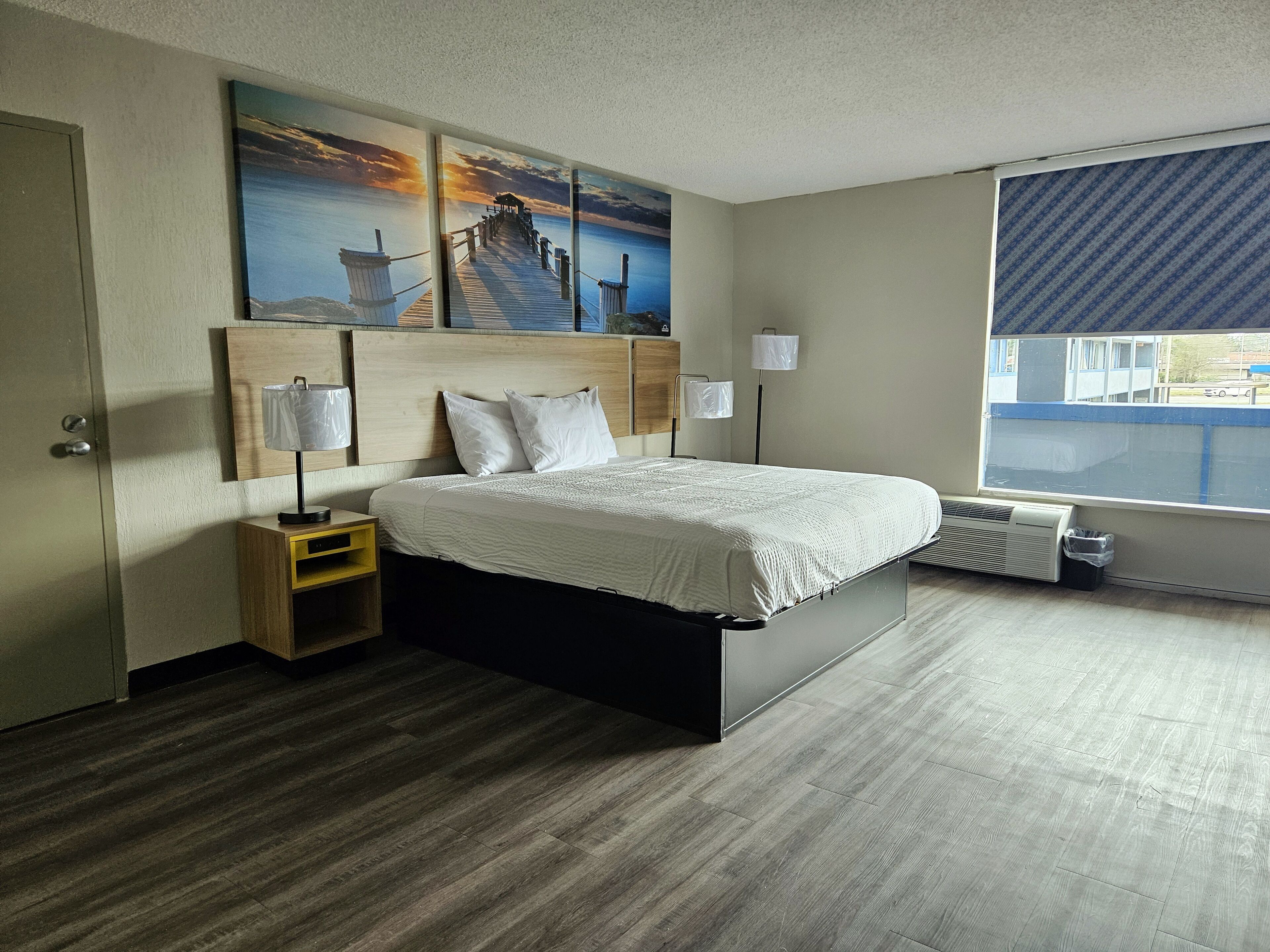 A hotel room with a bed, a nightstand, a lamp, a radiator, and a window with a view of the outside.