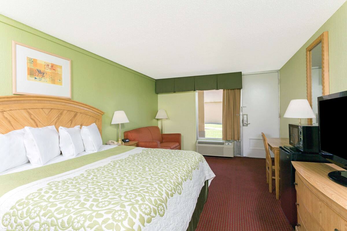 A hotel room with a bed, a desk with a television, a chair, a desk lamp, a bedside table, and a window with curtains.