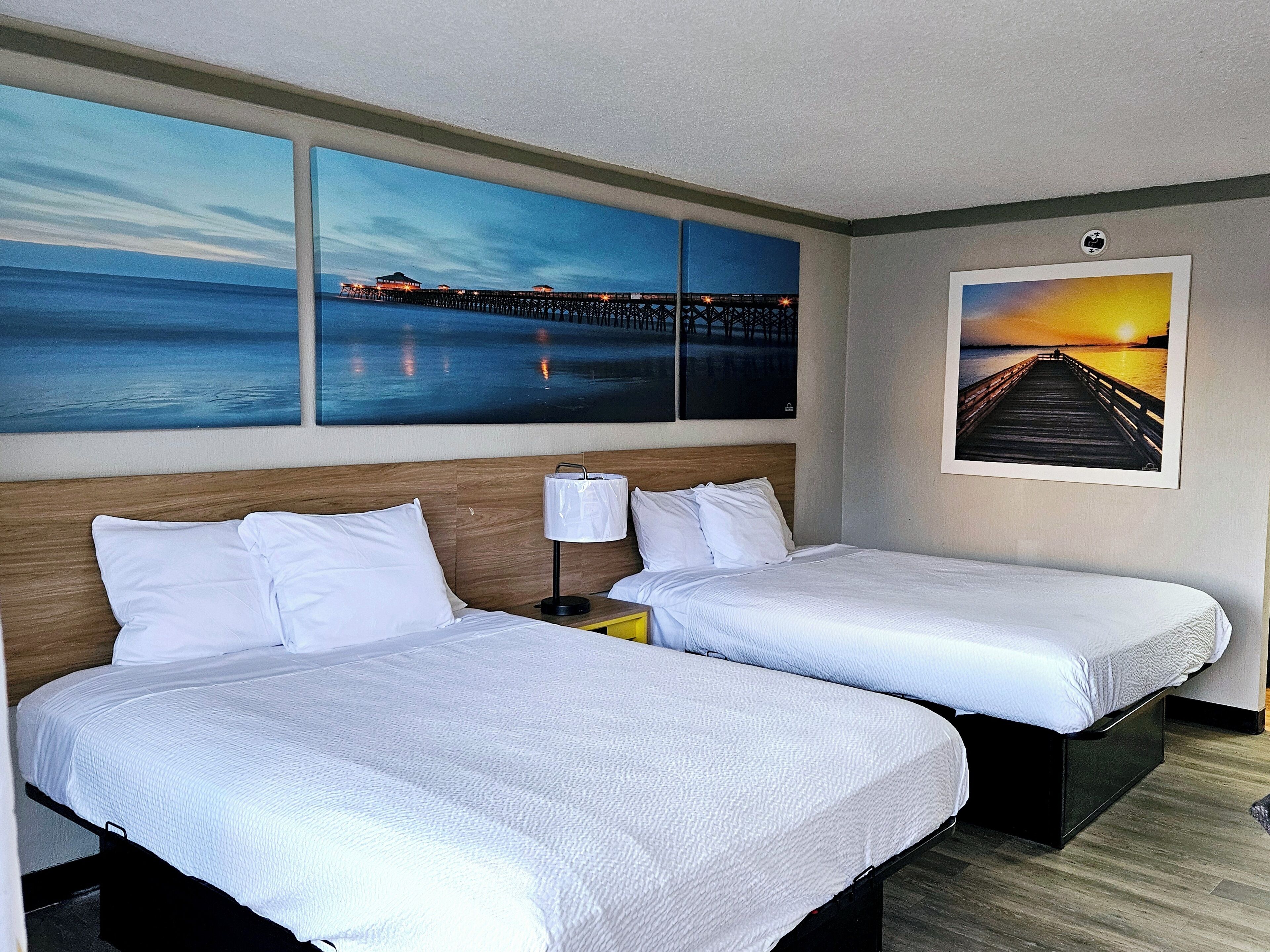 A hotel room with two single beds, a wooden headboard, a nightstand with a lamp, and artwork depicting a pier and sunset.