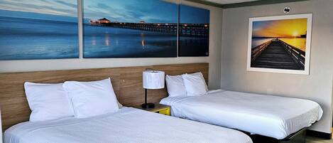 A hotel room with two single beds, a wooden headboard, a nightstand with a lamp, and artwork depicting a pier and sunset.