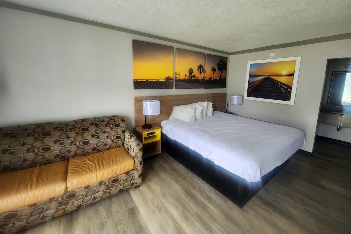 A hotel room with a bed, a sofa, two wall-mounted lamps, and two paintings depicting sunsets.