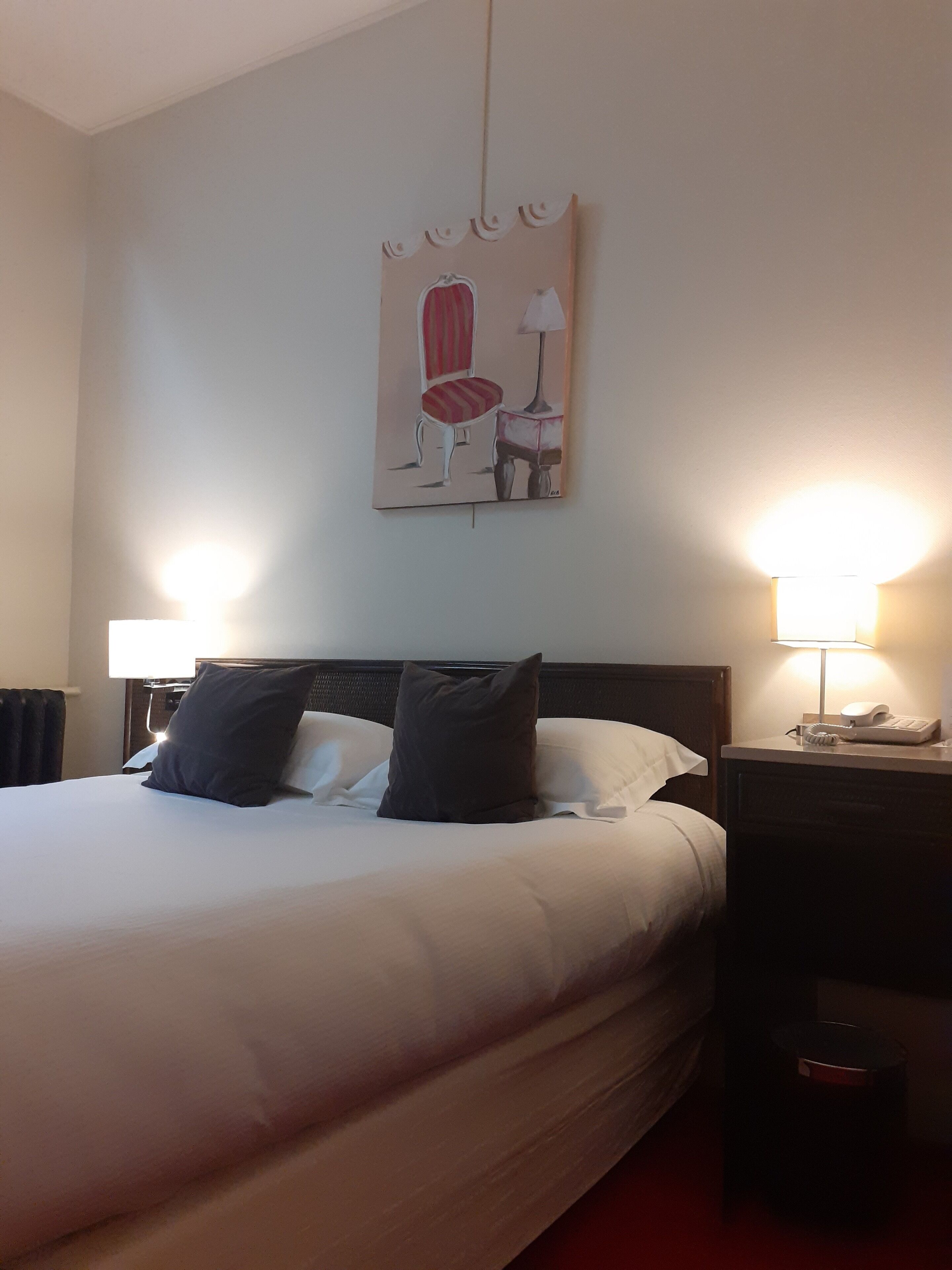 A hotel room with a large bed, two bedside lamps, a nightstand with a phone, and a painting on the wall.