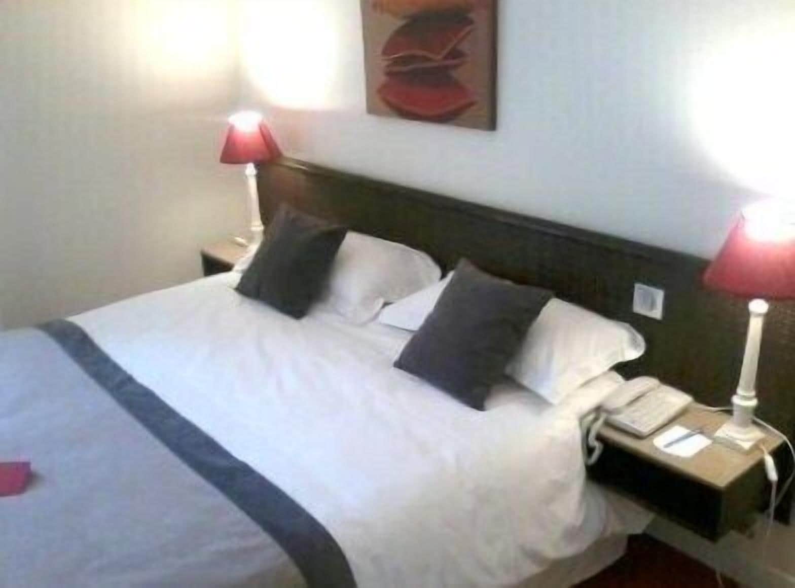 Quarto de hotel com uma cama, duas luminárias de cabeceira, um criado-mudo e um quadro na parede.