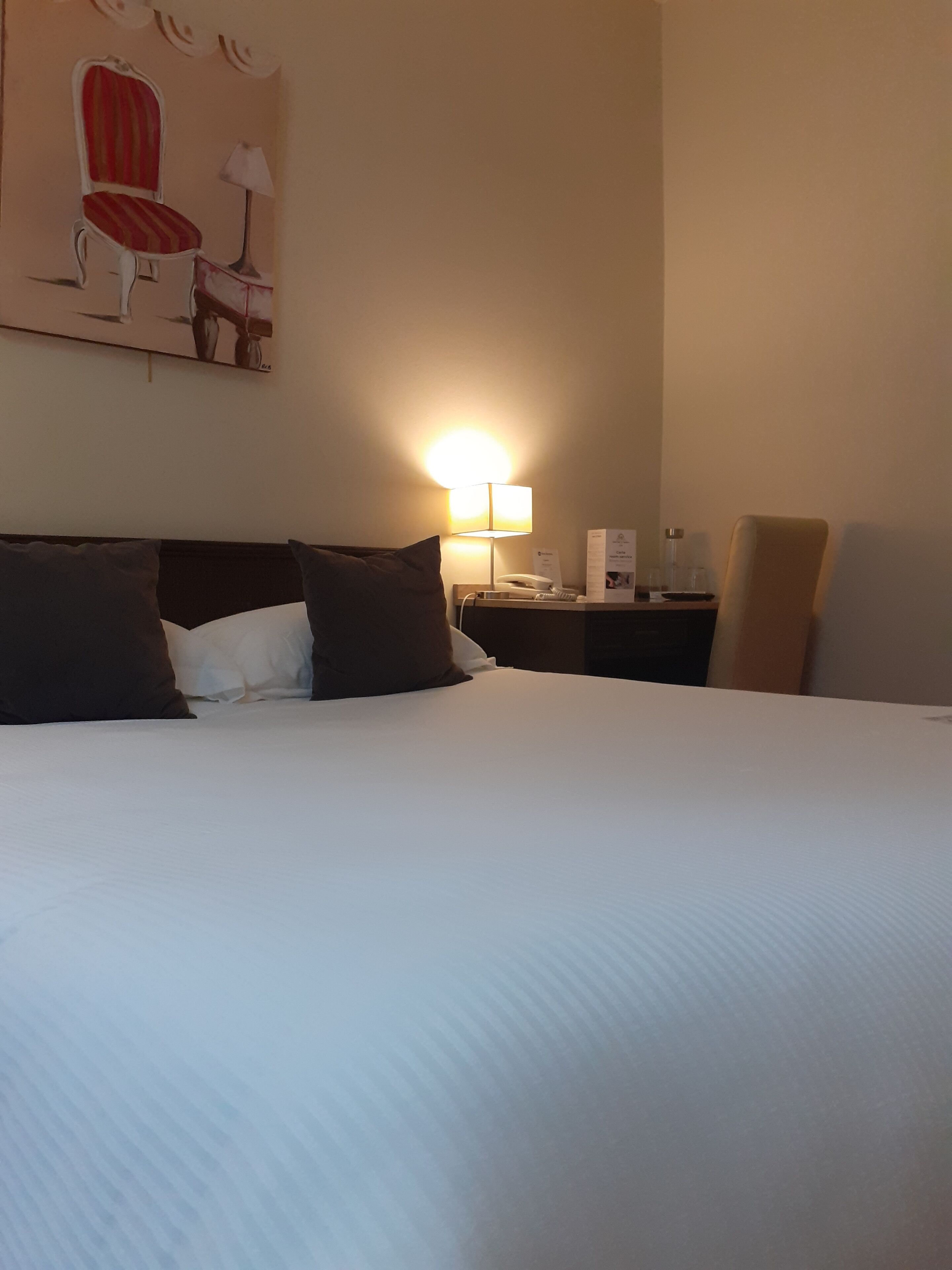 Ein Hotelzimmer mit einem großen Bett, einem Nachttisch mit Lampe, einem Sessel und einem Bild an der Wand.