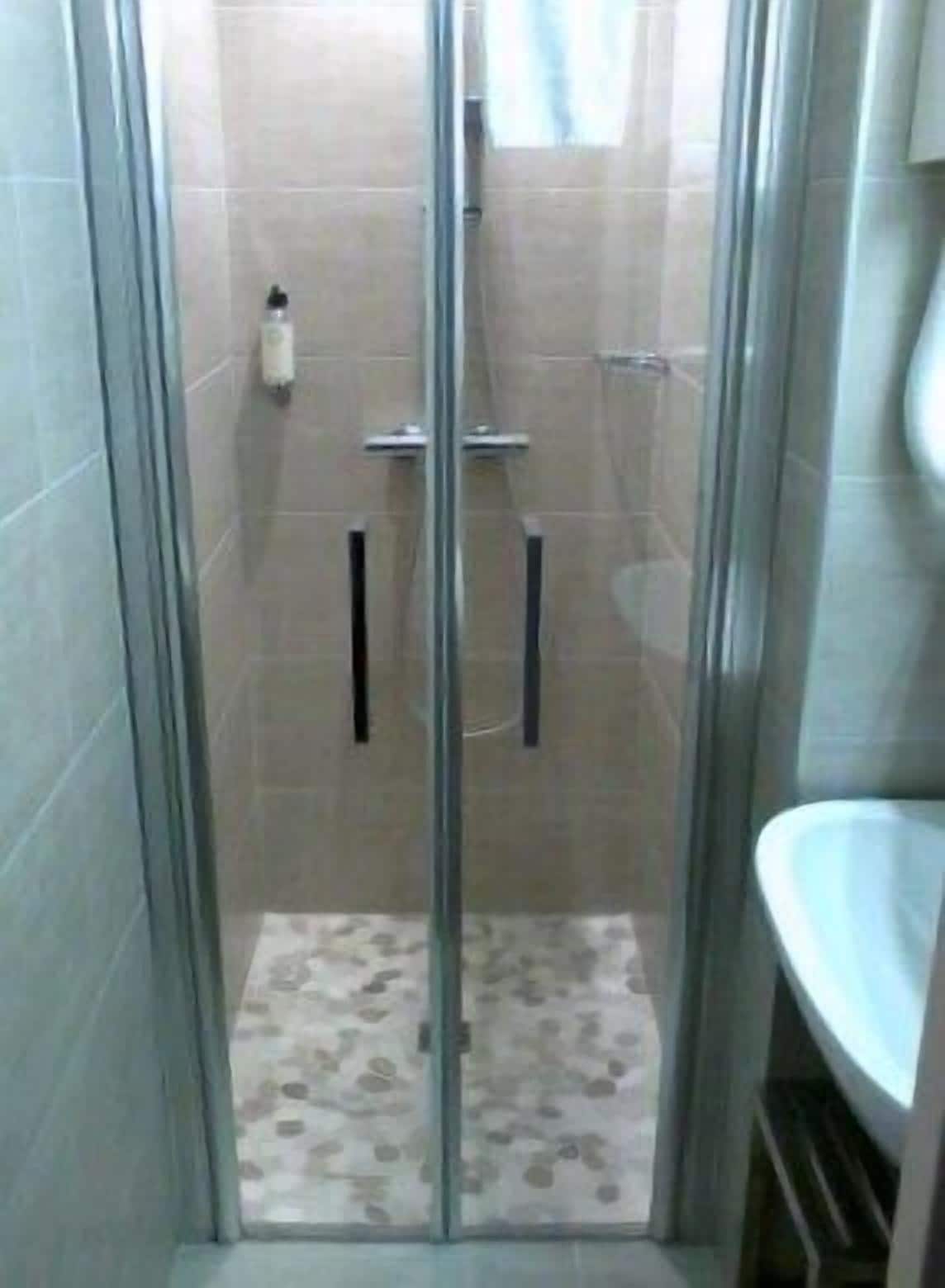 Une cabine de douche fermée par des parois de verre, avec un sol carrelé, une douchette à main et un flacon de shampoing fixé au mur.