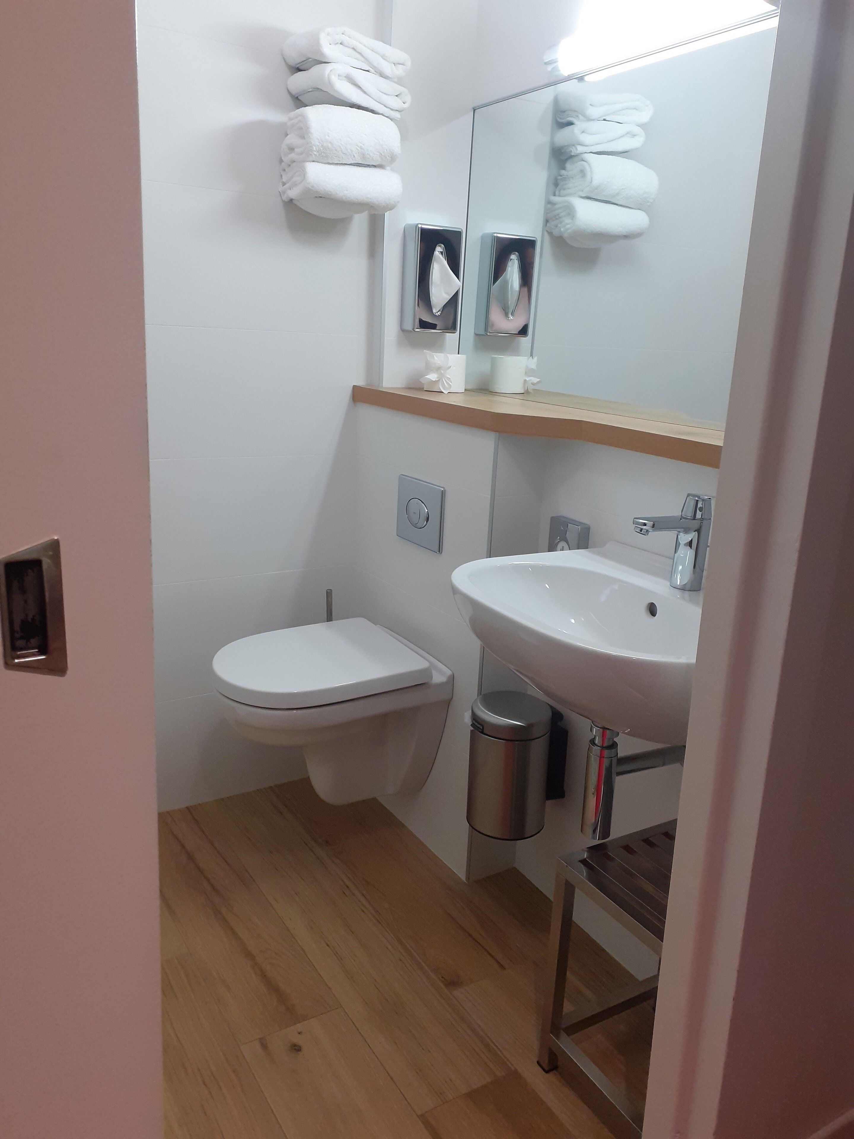 Ein Badezimmer mit Toilette, Waschbecken und Spiegel. Handtücher sind an der Wand gestapelt und ein Mülleimer befindet sich neben dem Waschbecken.
