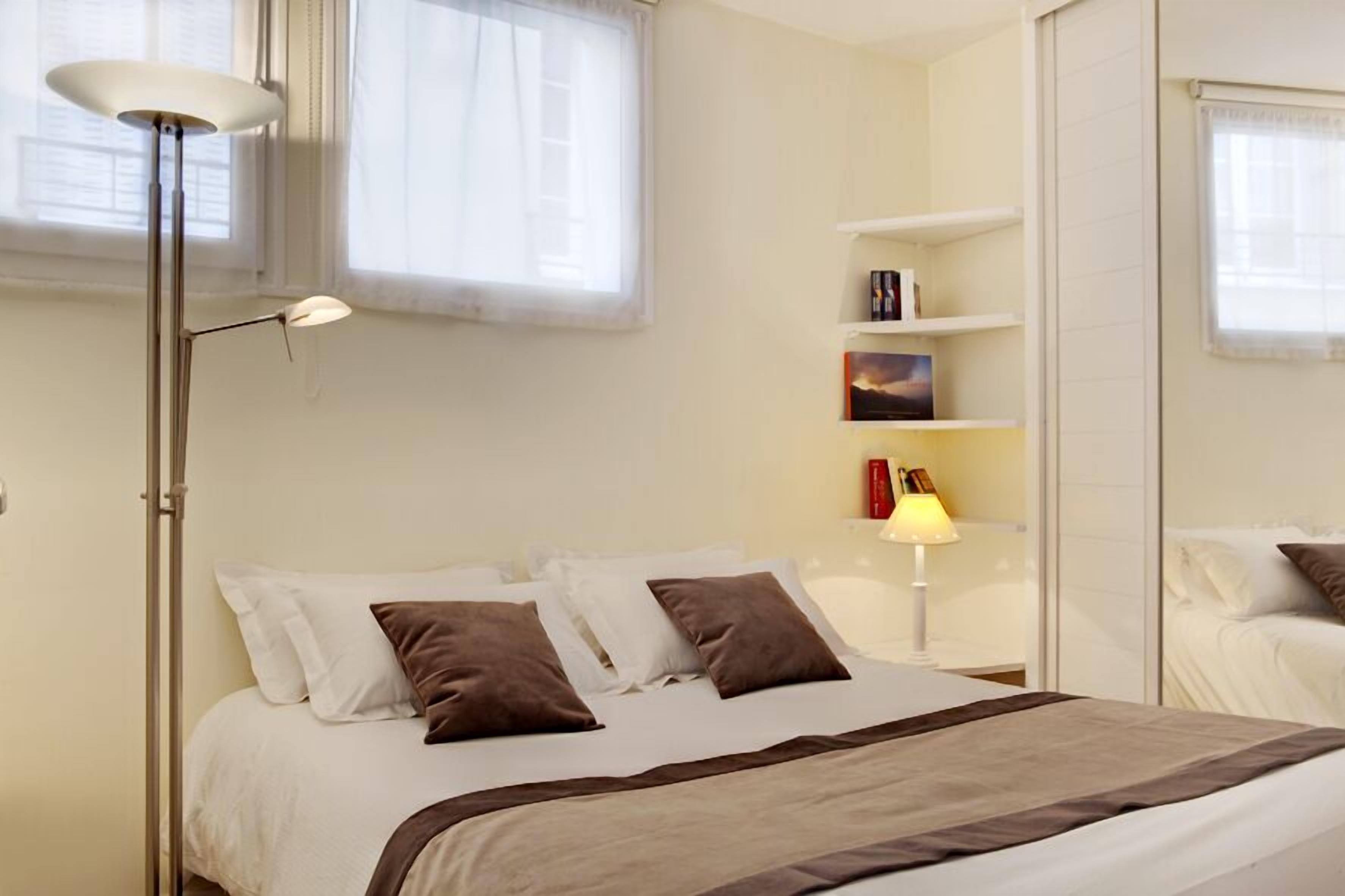 Una cama bien hecha con ropa de cama blanca y marrón, una lámpara de pie y una mesita de noche con lámpara.