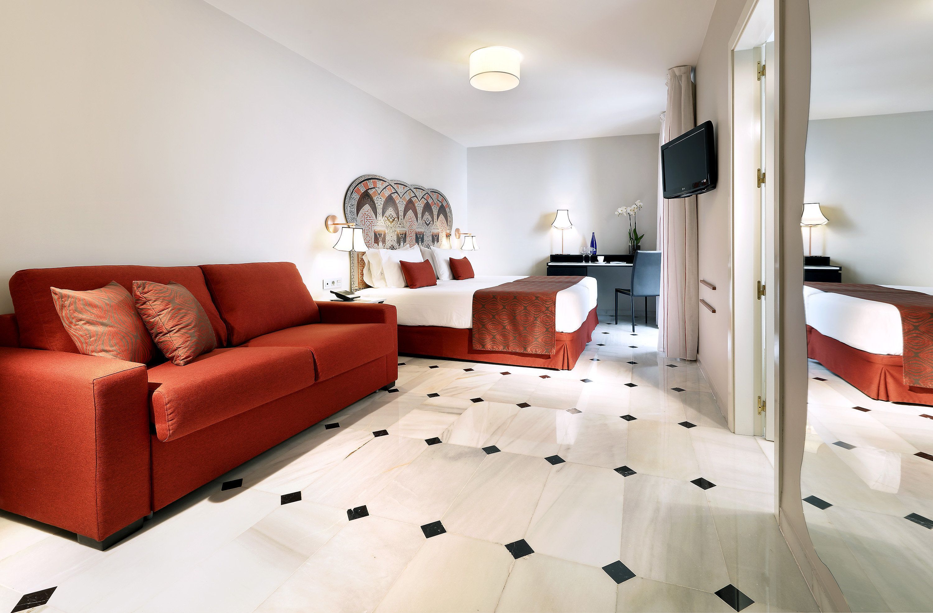 Une chambre d’hôtel moderne avec un canapé rouge, un grand lit doté d’une tête de lit à motifs, un téléviseur à écran plat et un coin repas comprenant une table et des chaises.