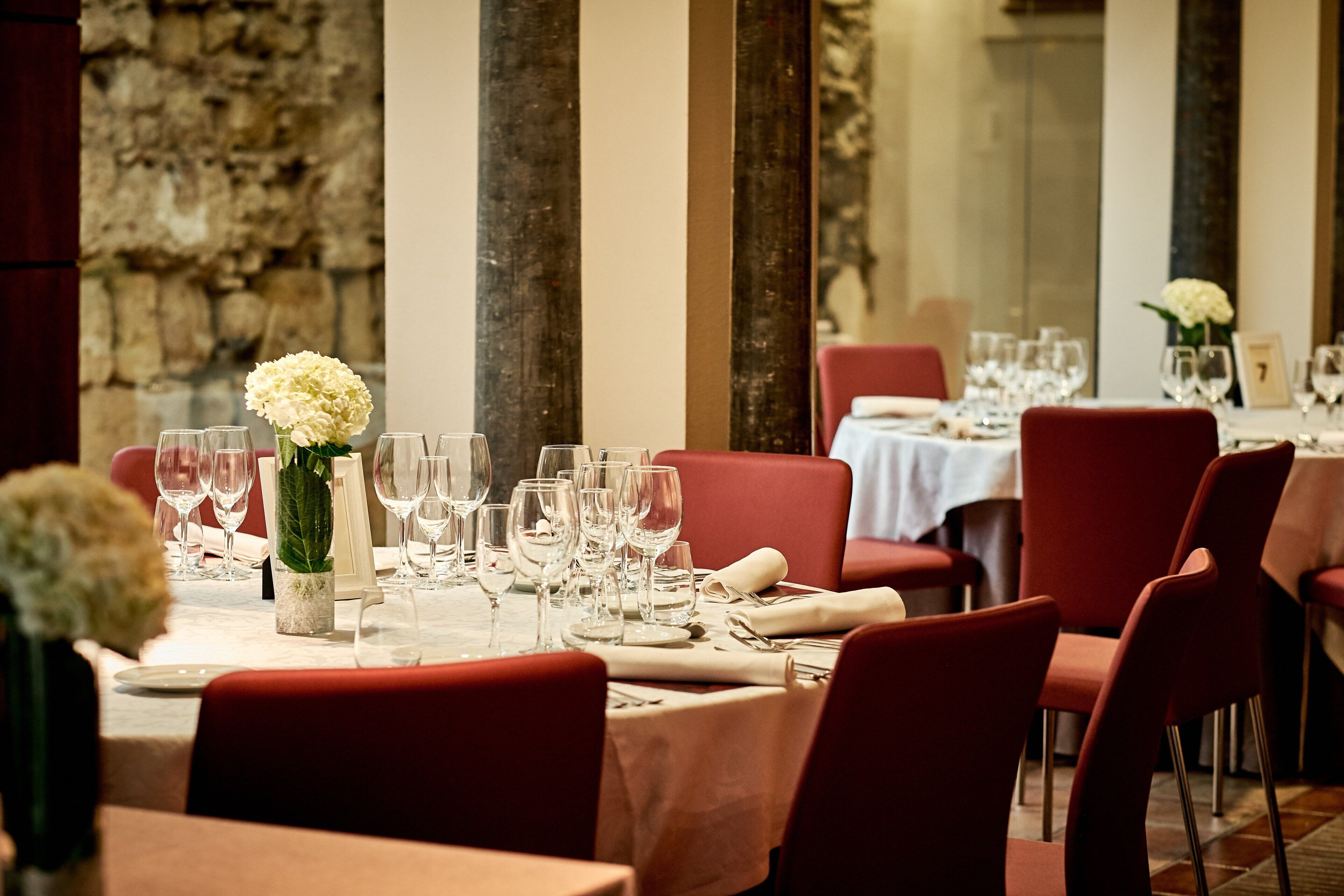 A restaurant with red upholstered chairs, white tablecloths, and a stone wall in the background.
