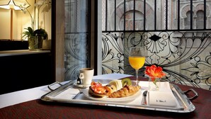 A breakfast tray with croissants, a muffin, and a glass of orange juice, a cup of coffee, and a vase with flowers on a table in a room with ornate metalwork and a view of a building with arches.