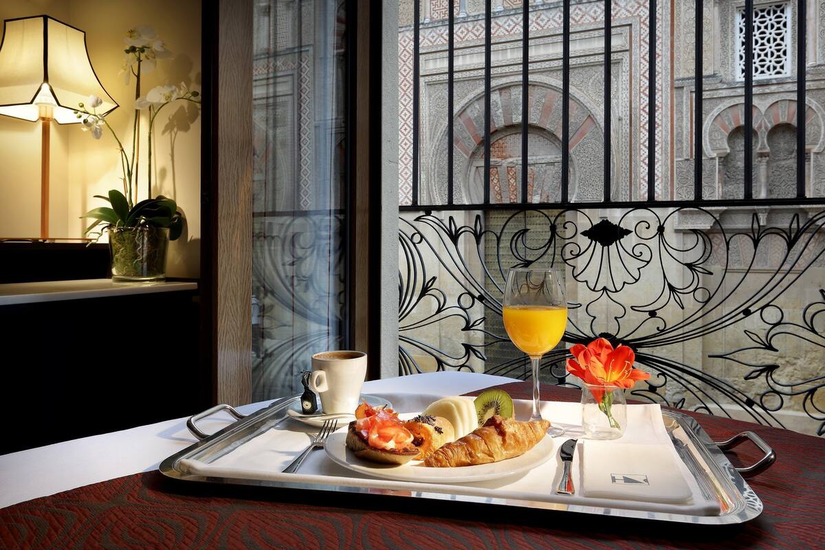 Un plateau de petit-déjeuner avec des croissants, un muffin et un verre de jus d’orange, une tasse de café et un vase de fleurs, le tout posé sur une table dans une pièce ornée de ferronnerie et offrant une vue sur un bâtiment à arcades.