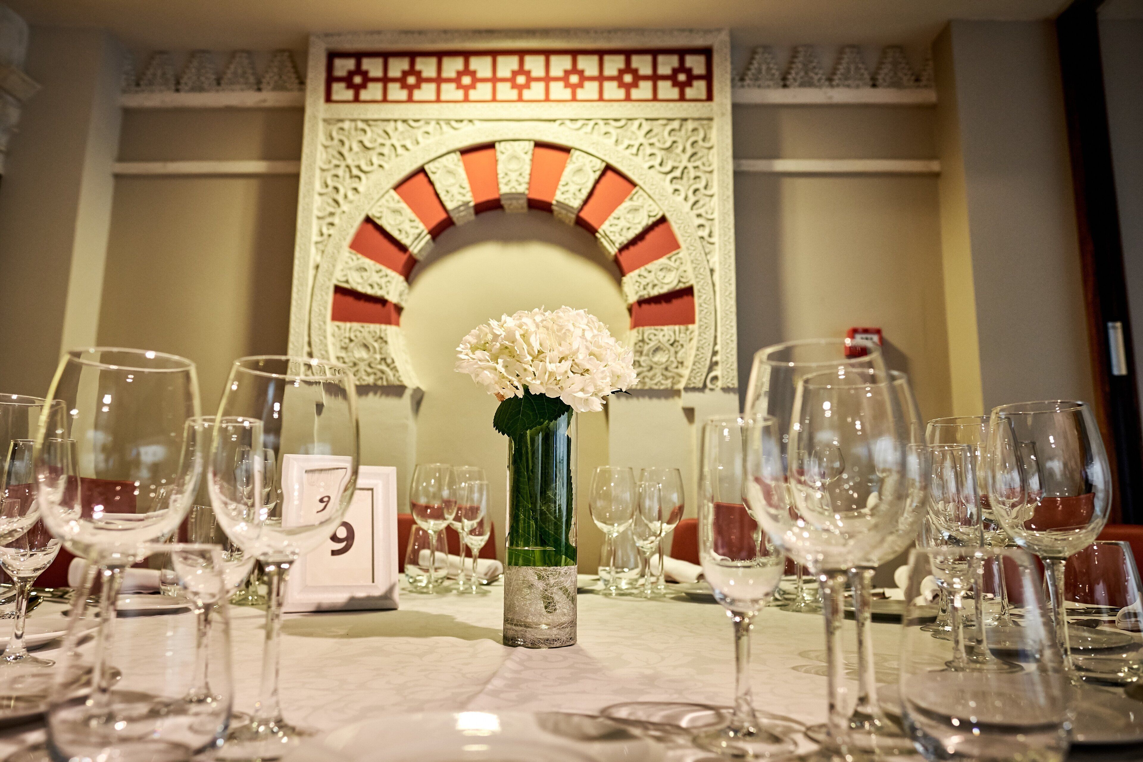 Une table dressée pour un dîner formel, avec des verres à vin, un vase de fleurs et une arche décorative sur le mur.