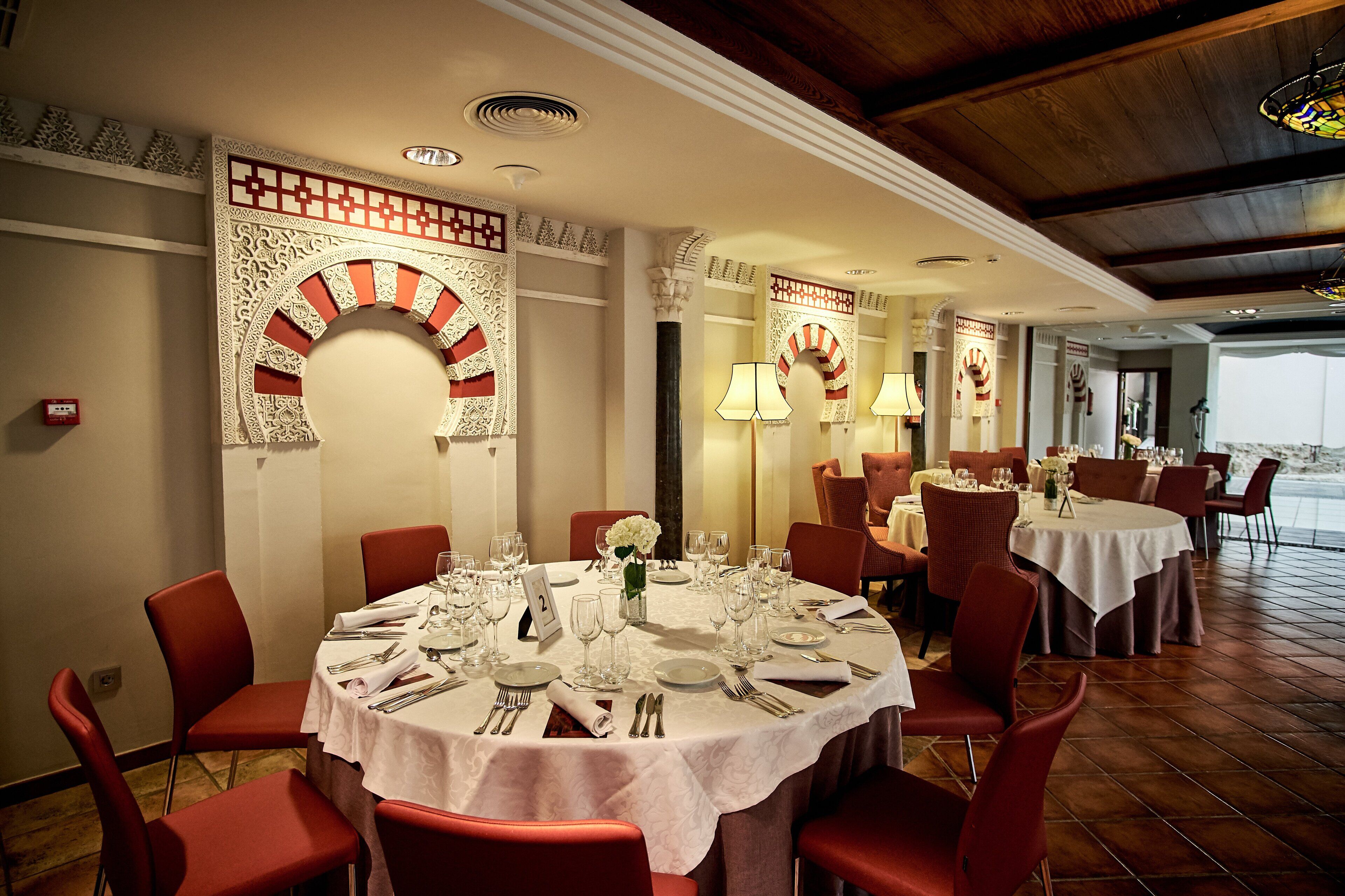 Un comedor con mesas redondas dispuestas para cuatro personas, sillas rojas y decoraciones ornamentales en las paredes.