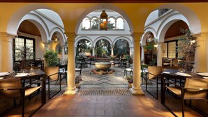 A spacious outdoor dining area with multiple tables and chairs, surrounded by arches and columns, featuring a central fountain and potted plants.