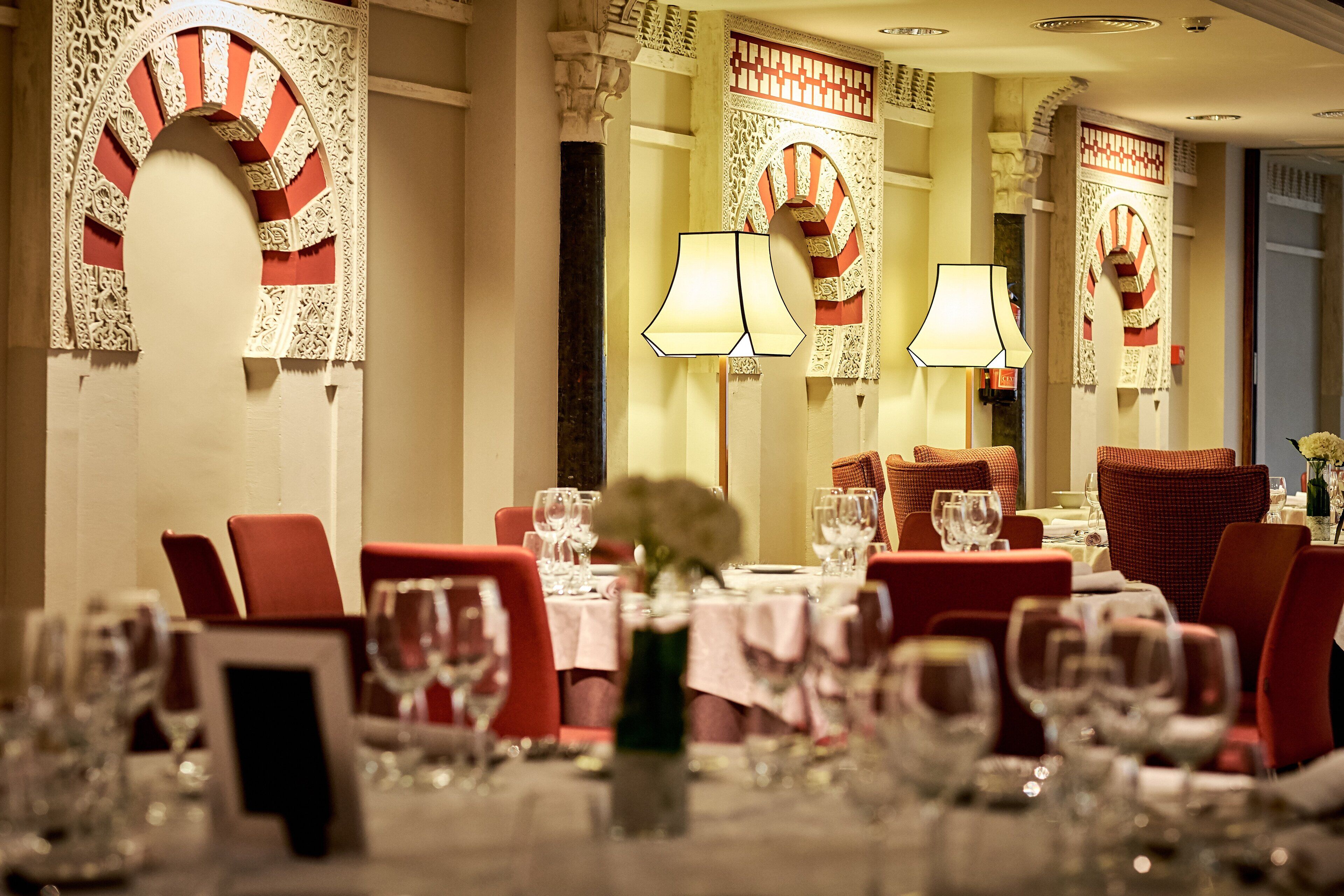 A dining area with red chairs, tables set with wine glasses, and decorative arches on the walls.