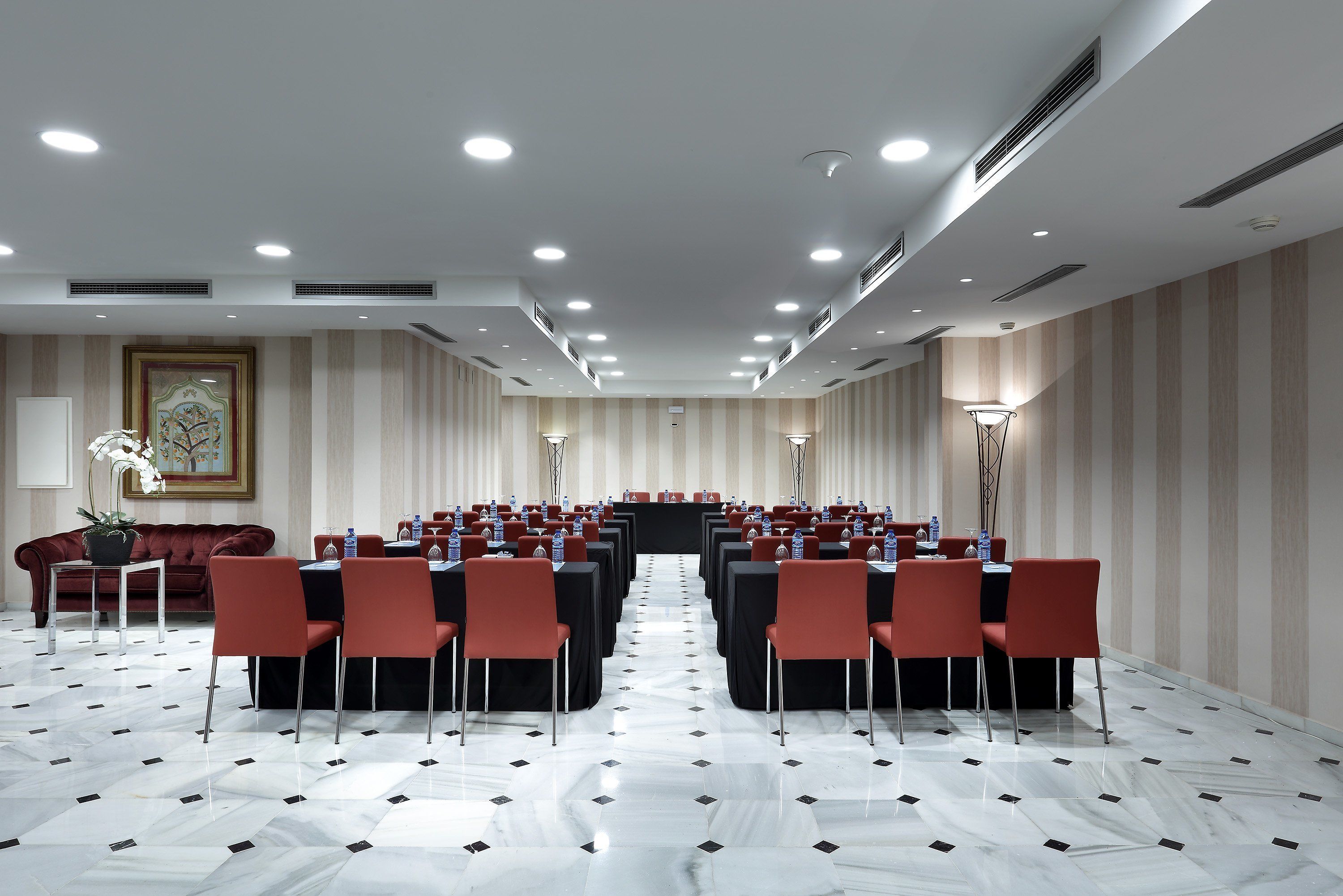 Un salón de conferencias con filas de mesas y sillas, un sofá rojo y un cuadro grande en la pared.