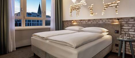 Un lit double dans une chambre d’hôtel, avec un mur en briques et une vue sur la ville par la fenêtre.