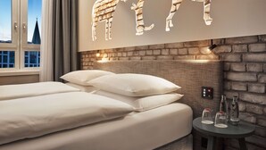 A hotel room with a brick wall, a bed with white linens, a small table with bottles, and a window with curtains. - H+ Hotel Bremen (Bremen)