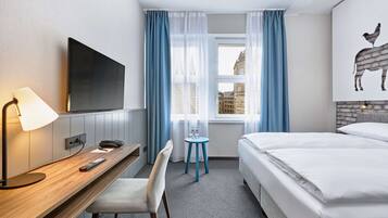 Ein Hotelzimmer mit einem Bett, einem Schreibtisch mit Lampe, einem Sessel, einem Fernseher und einem Fenster mit Vorhängen.