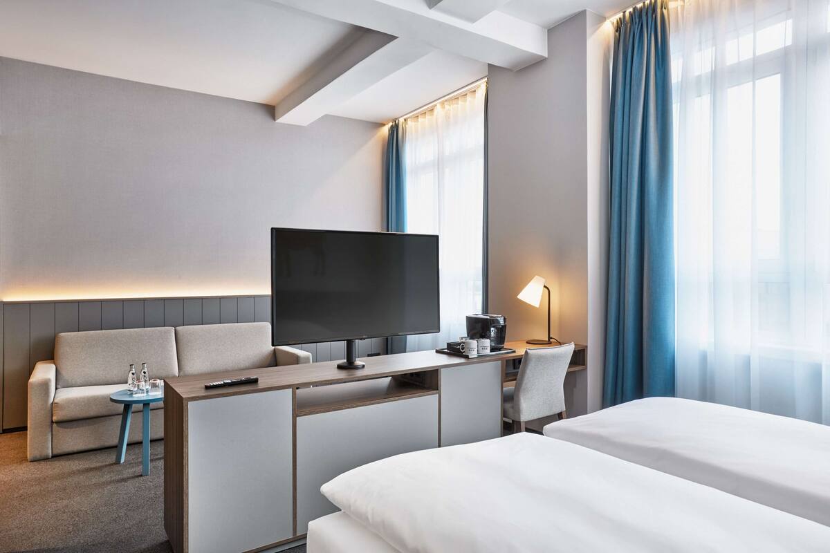 A modern hotel room with a large bed, a sofa, a desk with a flat-screen TV, a coffee machine, and a lamp.