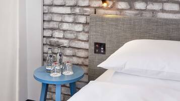 Ein Hotelzimmer mit Backsteinwand, einem Bett mit weißen Leinen, einem blauen Tisch mit Flaschen und einer Steuerung am Kopfteil.