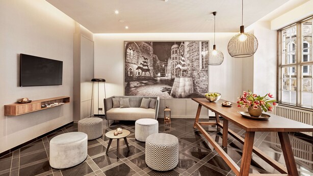 A modern living room with a large wall-mounted TV, a sofa, ottomans, a dining table with a vase of flowers, and a large framed picture of a street scene. - H+ Hotel Bremen (Bremen)