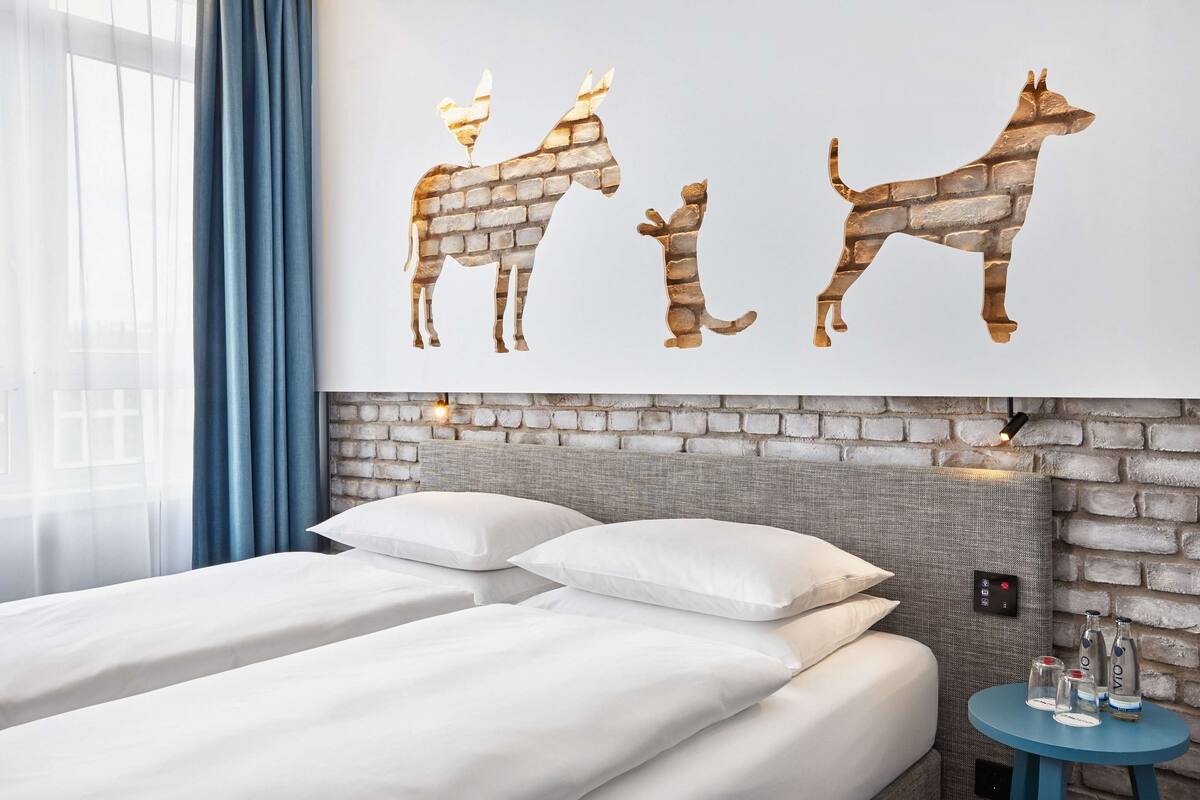 A hotel room with two beds, a brick-patterned headboard, a wall art of animals, and a small table with bottles.