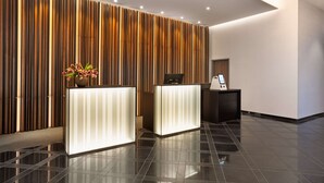 A modern reception area with a wooden panel wall, two reception desks, a potted plant, and a digital information kiosk. - H+ Hotel Bremen (Bremen)