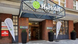 The exterior of a H+Hotel with a glass entrance, brick facade, and large potted plants. - H+ Hotel Bremen (Bremen)